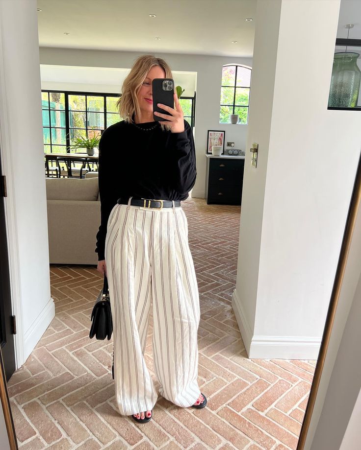 Mulher em um ambiente moderno, usando blusa preta e calças listradas brancas.